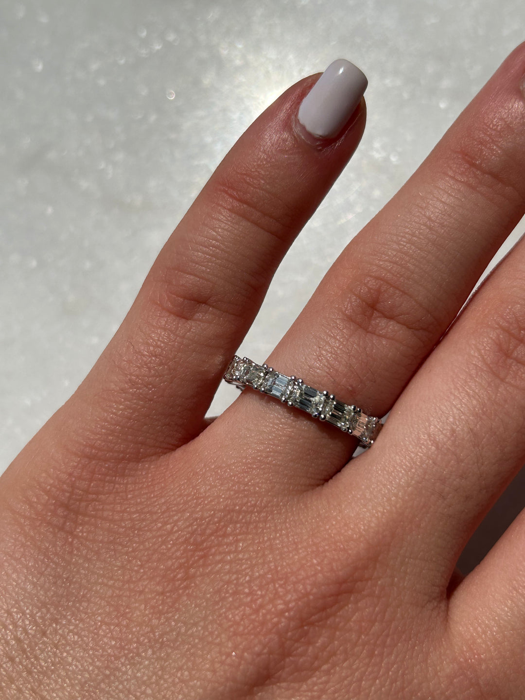 An image featuring a model elegantly displaying an emerald diamond eternity band, the sparkling emerald-cut diamonds intricately set around the band, catching the light with mesmerizing brilliance as the model's hand moves gracefully, showcasing the luxurious and timeless beauty of the jewelry piece against a subtle background.